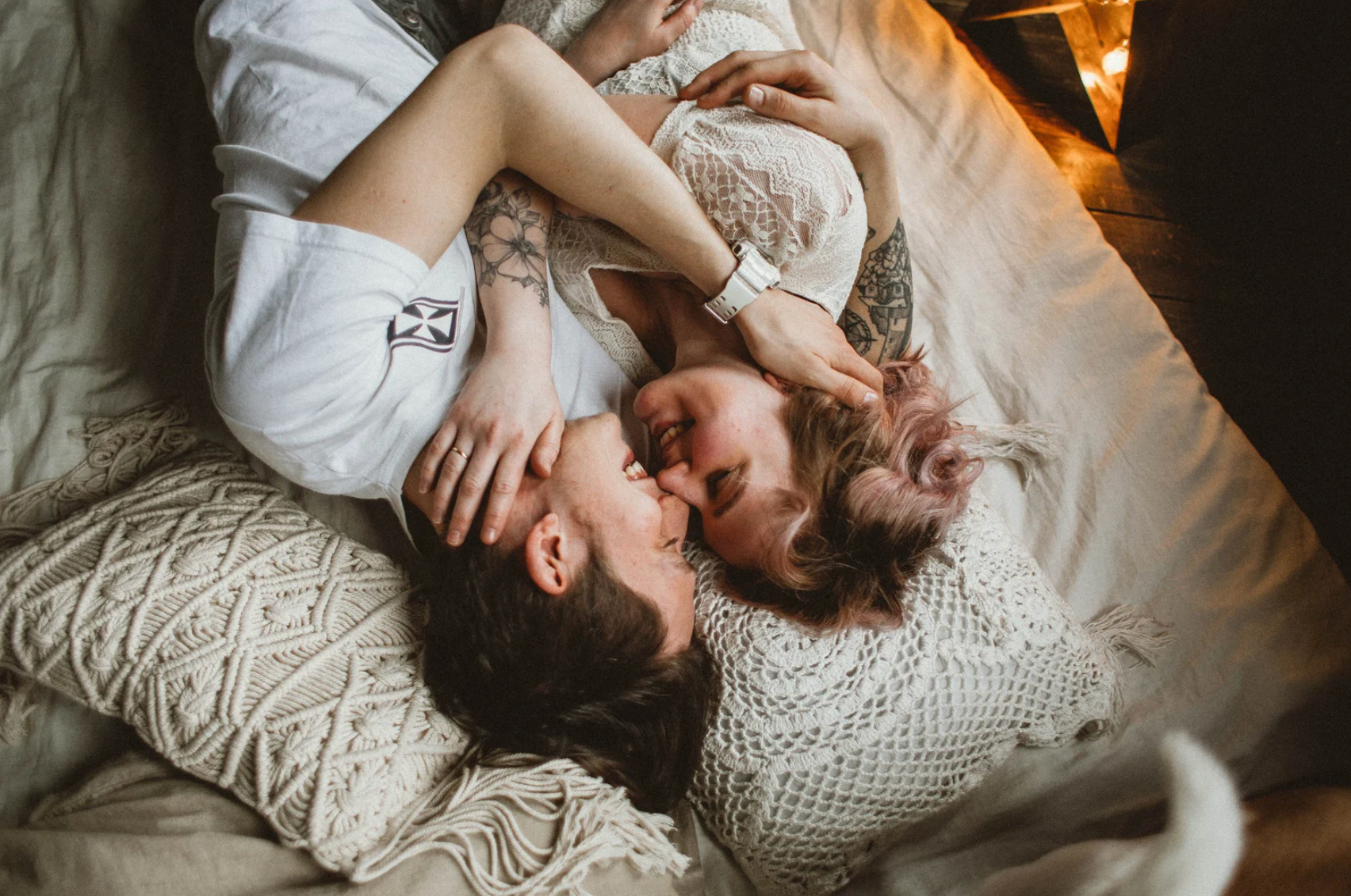 Couple allongé sur un lit, enlacé et souriant, dans une ambiance chaleureuse et intime, avec des coussins en macramé et une lumière douce.