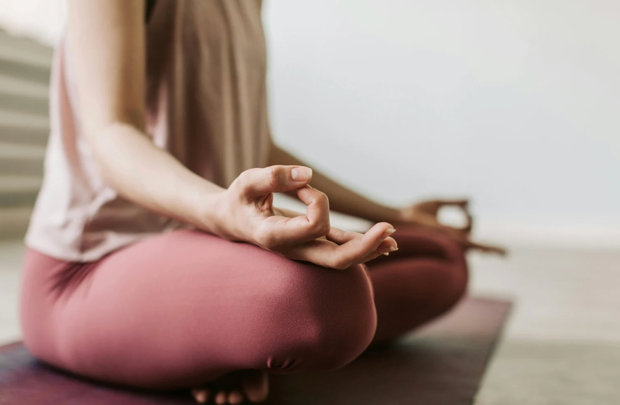 Personne assise en tailleur sur un tapis de yoga, main posée sur le genou en geste de méditation, dans une ambiance calme et épurée.