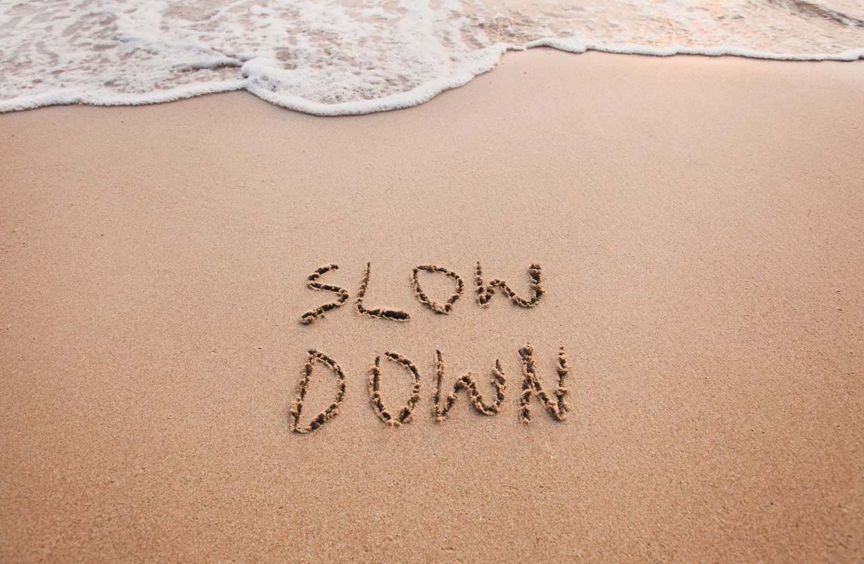 Vue d’une plage de sable avec la mer en haut de l’image et les mots « SLOW DOWN » écrits dans le sable.