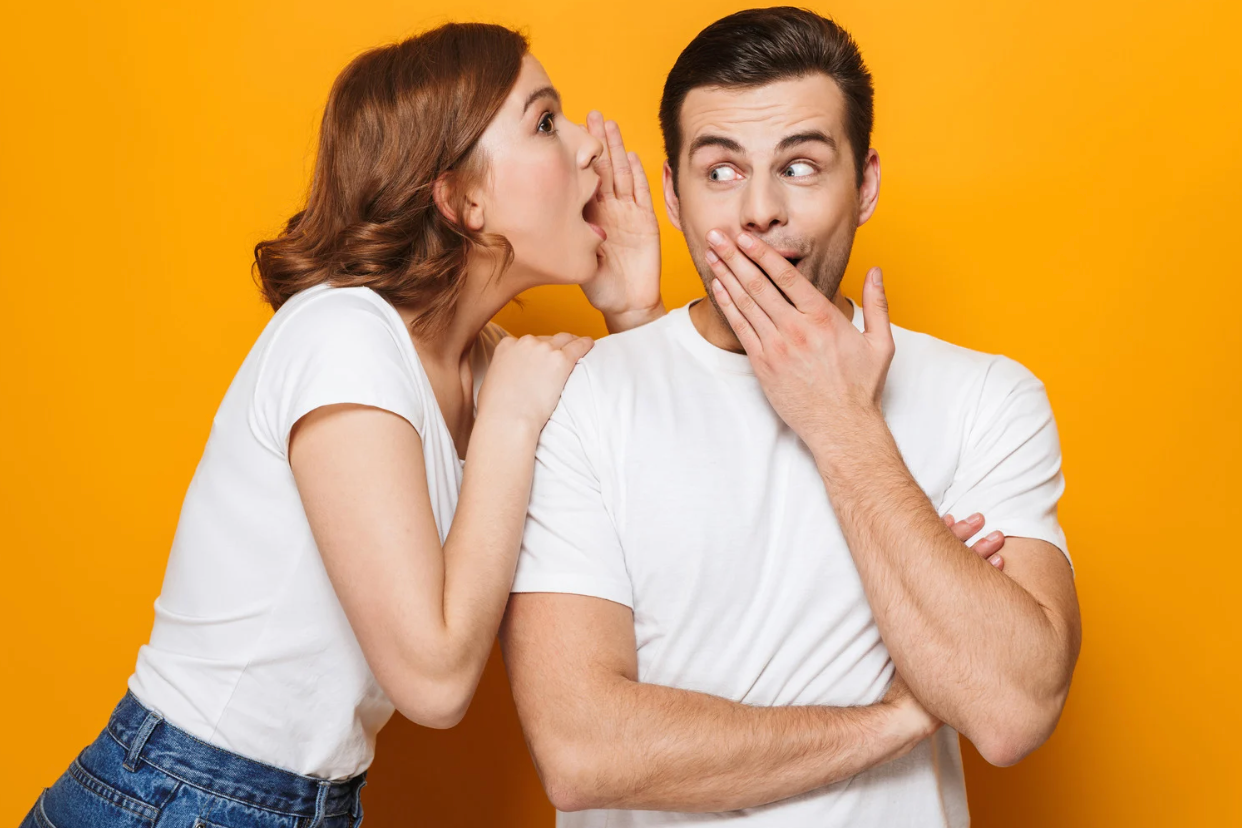 Femme souriante chuchotant un secret à l’oreille d’un homme surpris, tous deux en t-shirt blanc devant un fond orange uni.